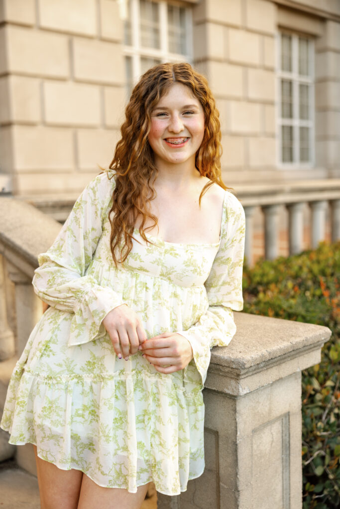 Senior girl with curly hair leaning on a stone railing and smiling, individual portrait from a senior friend photos session in Yucaipa by Kaitlyn Dawn Photography.