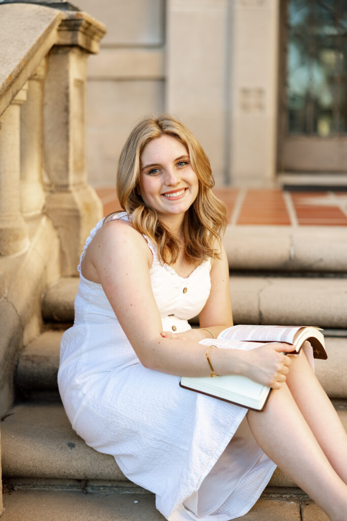 Senior girl in Redlands sitting on steps holding an open book, wearing a white dress and smiling naturally during her senior photos.