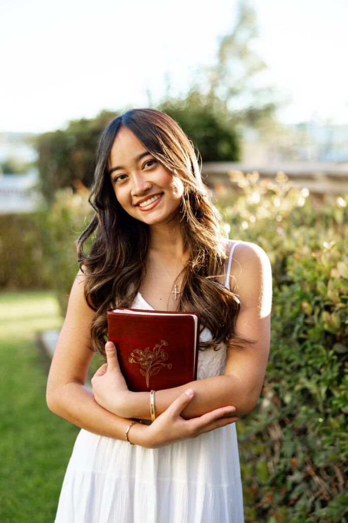 Senior girl in white dress holding a journal and smiling outdoors in Redlands, illustrating indoor vs outdoor senior photos by Kaitlyn Dawn Photography.