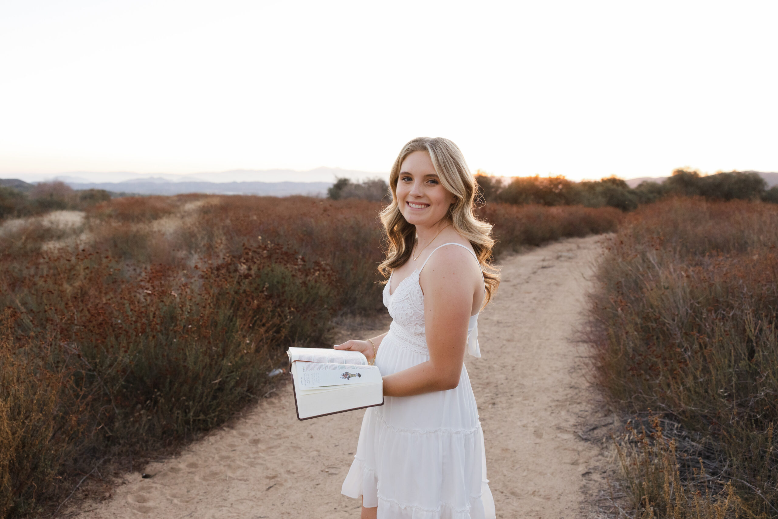 Yucaipa senior in a white dress holding a book outdoors, a personalized senior photo prep idea for meaningful portraits.