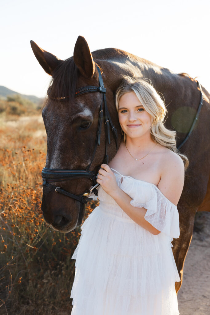 High school girl in a white dress standing beside her horse in a field at sunset, by Kaitlyn Dawn Photography.
