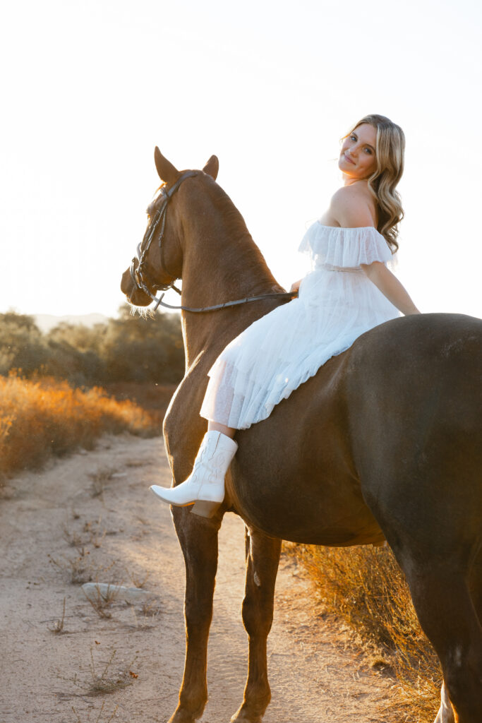 High school girl in a white dress sitting on a horse at golden hour, scenic outdoor session by Kaitlyn Dawn Photography.