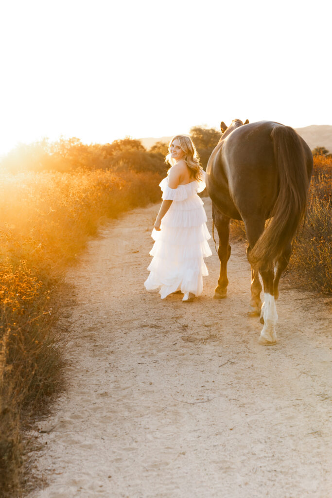 Cate in a flowing white dress walking alongside her horse on a sunlit trail, dreamy golden hour Redlands senior portrait story.