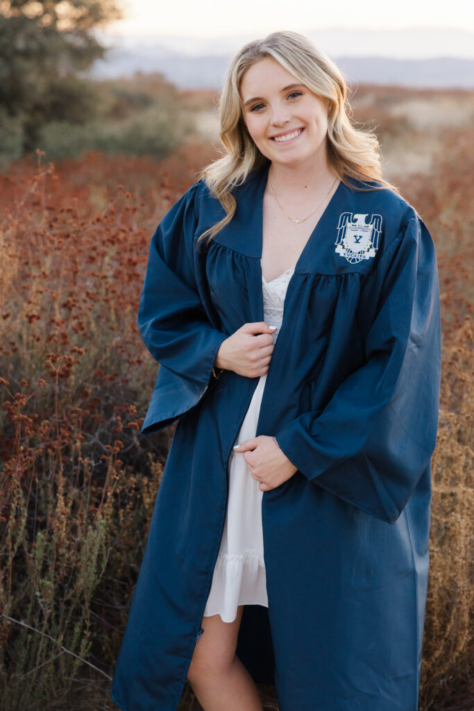 Close-up of senior girl in navy graduation gown smiling in a field at golden hour, Inland Empire senior photography by Kaitlyn Dawn Photography.