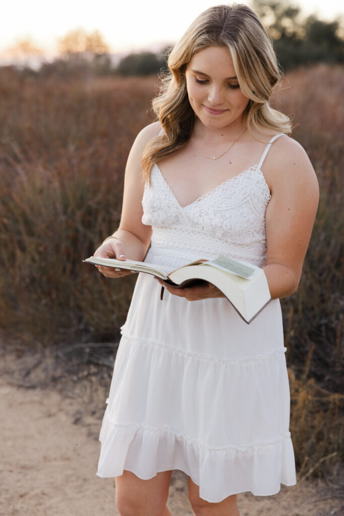 Girl in a white dress reading a book outdoors in a scenic field, soft sunset lighting, Inland Empire portraits by Kaitlyn Dawn Photography.