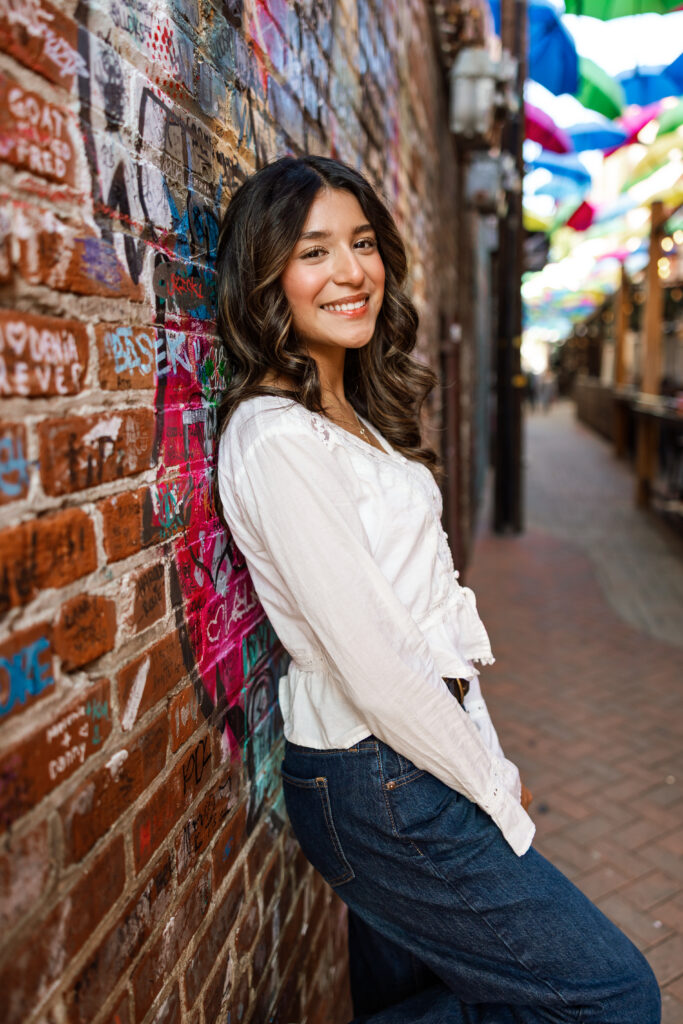 Senior leaning against colorful brick wall in white top and denim, highlighting best colors for senior photos with contrast and texture.