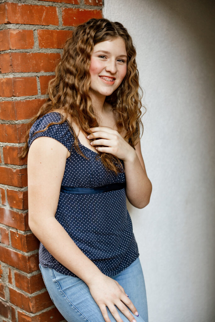 Senior wearing a navy patterned top with light-wash jeans for a casual portrait against a brick wall captured by a Yucaipa senior photographer.