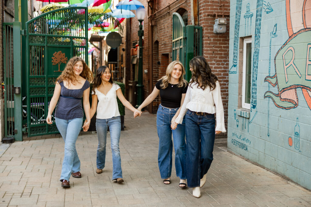 Four senior girls holding hands and walking through an urban alleyway, stylish senior friend photos in Yucaipa by Kaitlyn Dawn Photography.