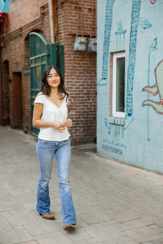 Senior walking downtown in a white top, flare jeans, and brown boots for a relaxed senior portrait outfit.