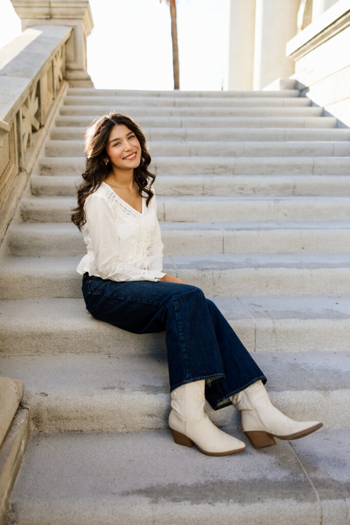High school senior sitting on outdoor steps in Redlands, wearing a white blouse and jeans, smiling during a casual portrait.