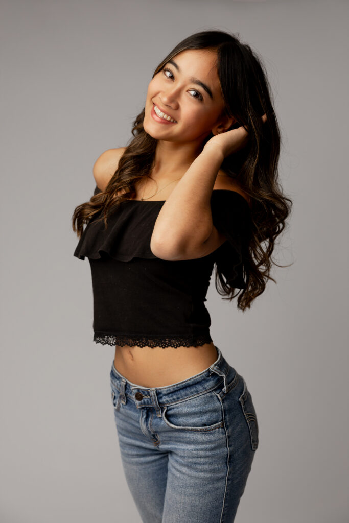 Studio senior portrait of girl smiling in black off-shoulder top and jeans in Redlands, highlighting indoor vs outdoor senior photos by Kaitlyn Dawn Photography.