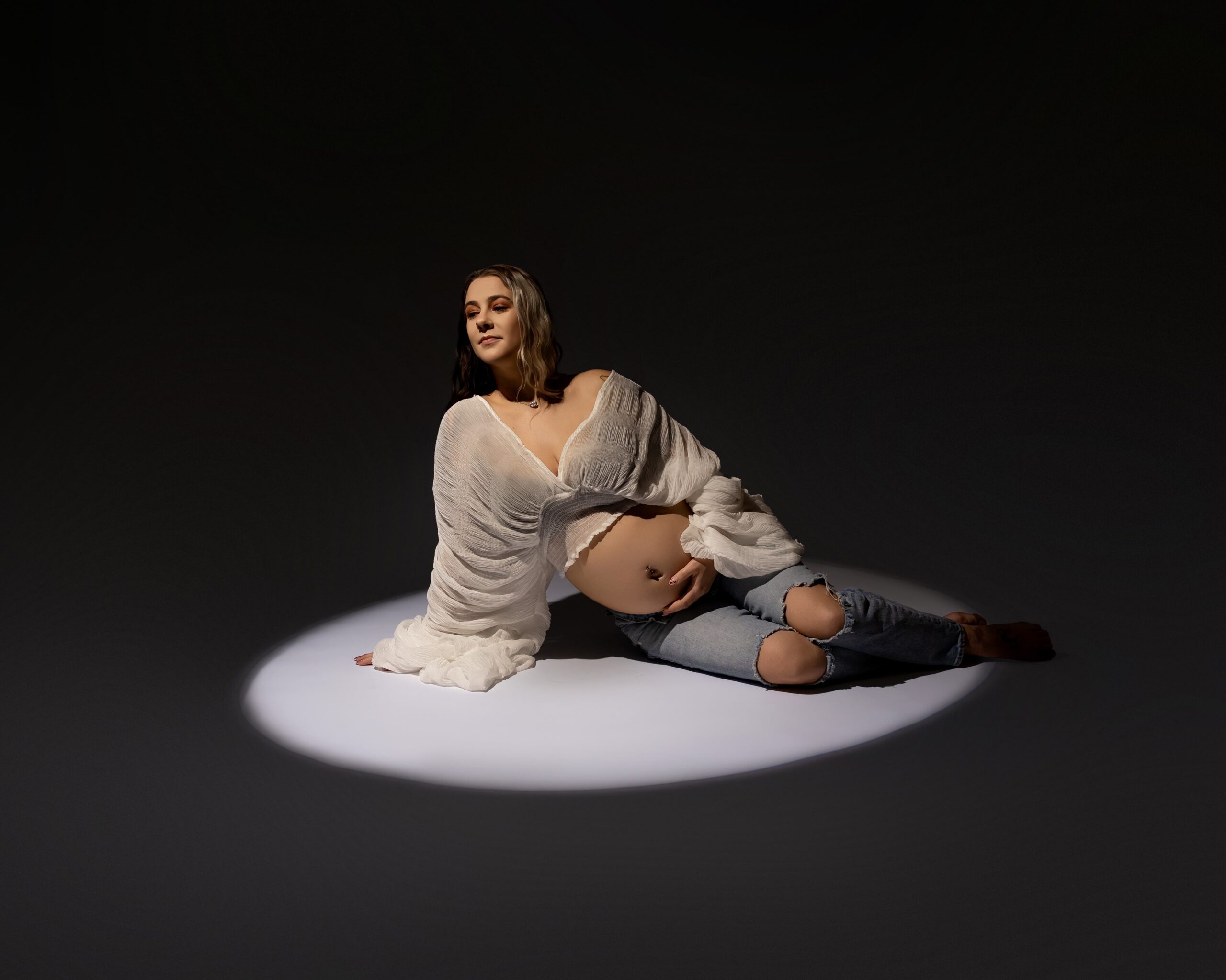 Pregnant mother sitting under dramatic studio lighting during maternity session, reflecting on the hardest week of pregnancy. Photograph by Yucaipa maternity photographer Kaitlyn Dawn Photography.