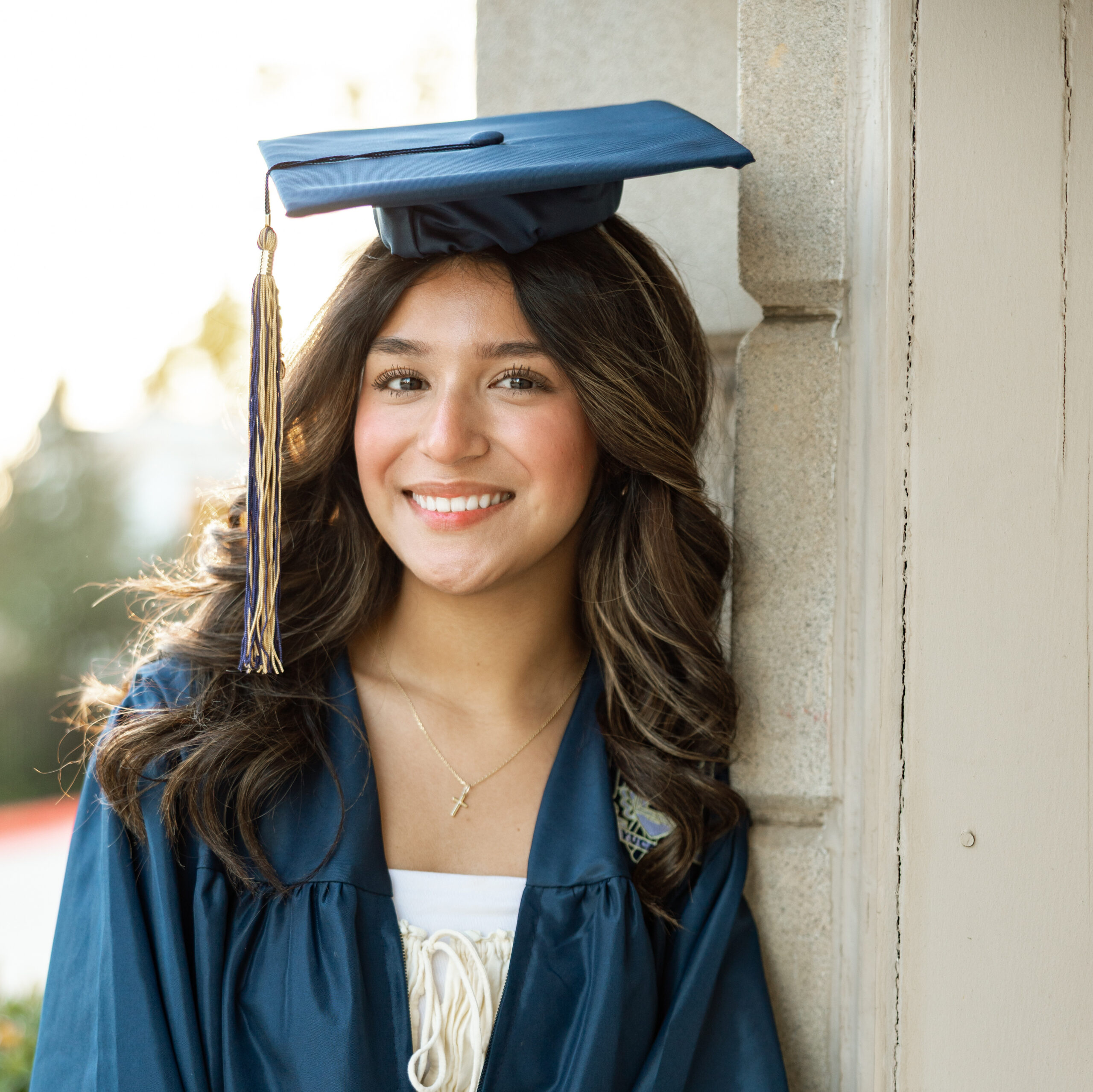 Smiling high school graduate in cap and gown leaning against a wall, capturing a warm and natural senior portrait experience in the Inland Empire by Kaitlyn Dawn Photography.