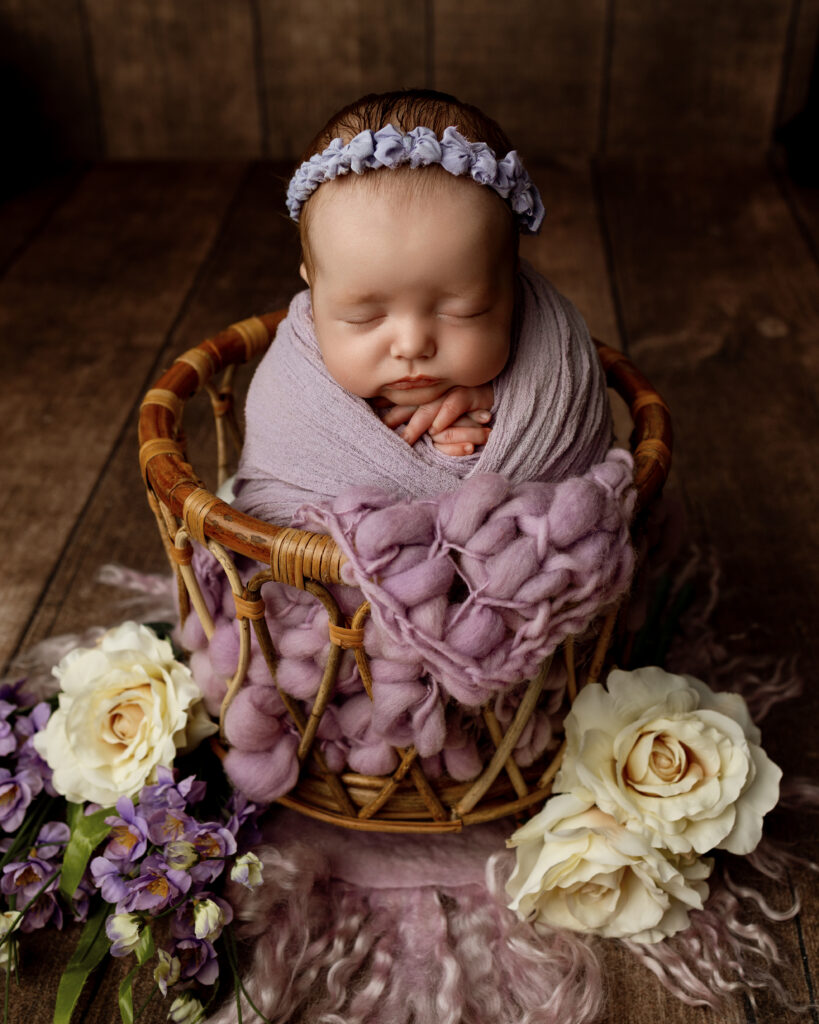 Sleeping newborn wrapped in a lavender swaddle in a wicker basket with purple flowers by an Inland Empire newborn photographer.