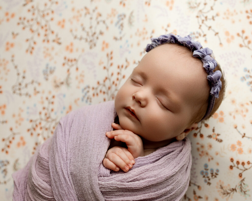 Peaceful newborn wrapped in a lavender swaddle with a headband on a patterned backdrop, celebrating 2 month old baby milestones and early baby development.