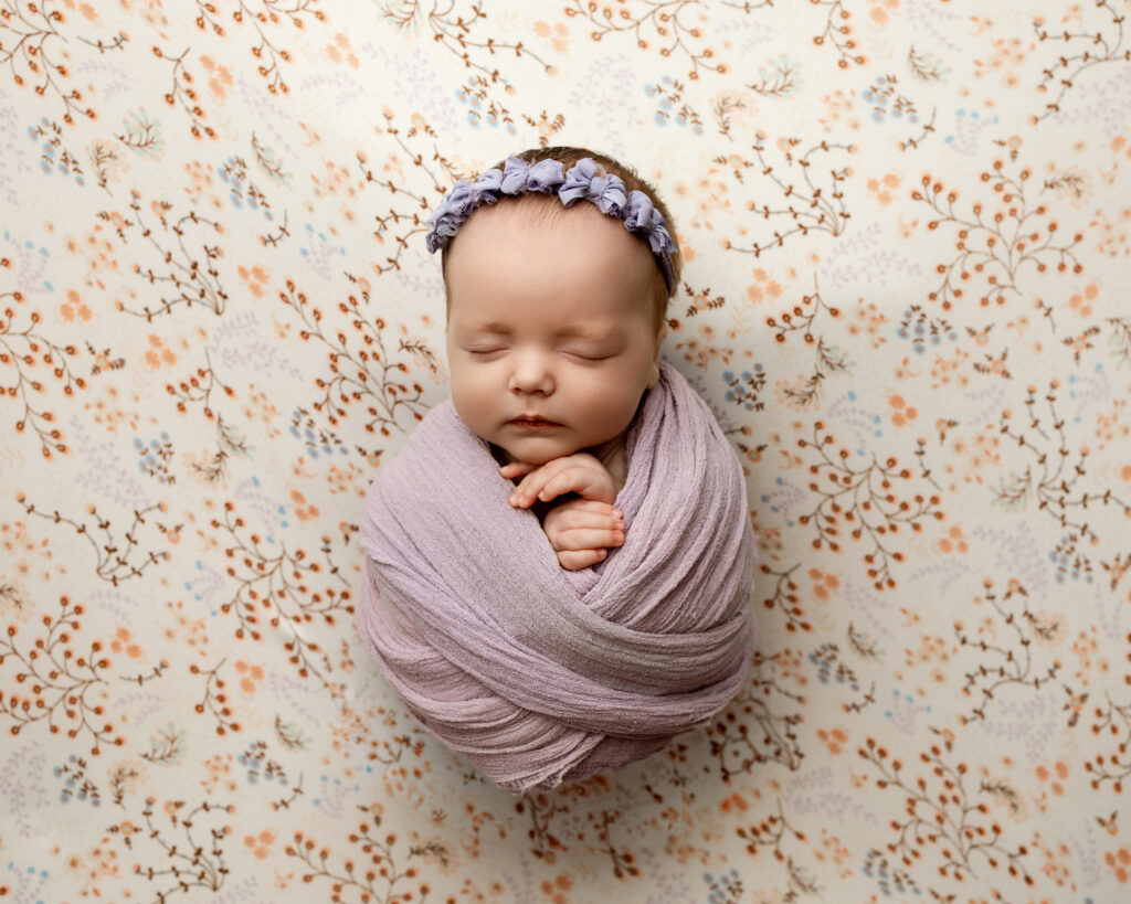 2 month old baby milestones featuring a sleeping newborn wrapped in a soft lavender swaddle on a delicate floral backdrop during a studio portrait session.
