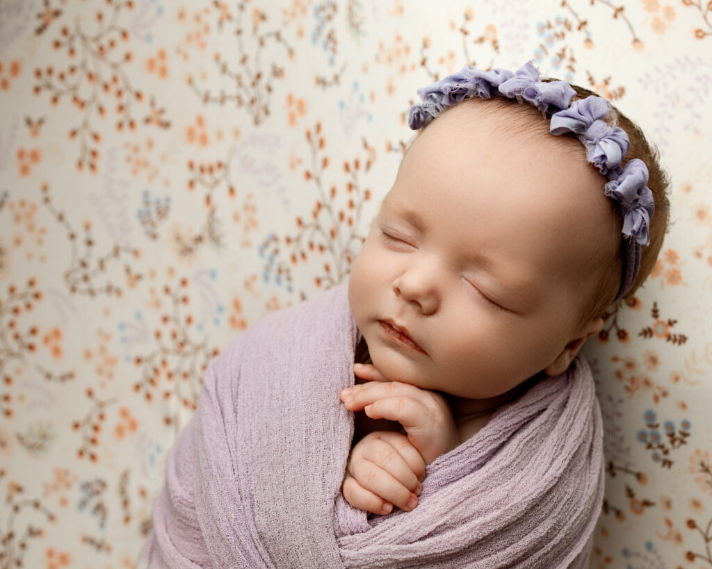 Close-up of a sleeping newborn in a lavender swaddle on a delicate floral backdrop by an Inland Empire newborn photographer.