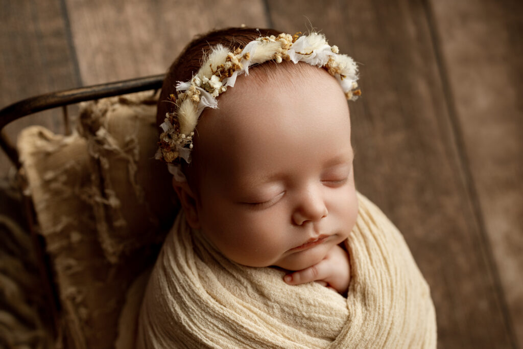 Newborn sleeping peacefully in a cream swaddle with a floral headband, photographed by a newborn photographer in the Inland Empire.