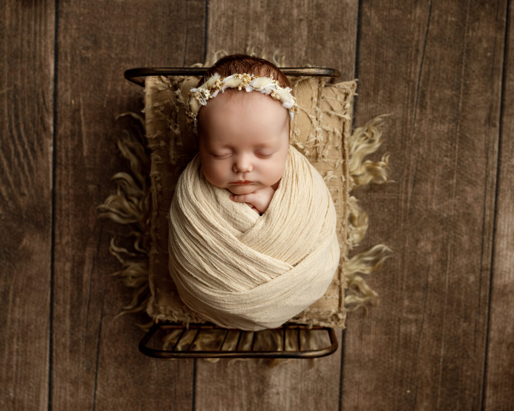 Newborn sleeping peacefully in a cream swaddle and floral headband in a vintage bed, a beautiful image of a 2 month old baby captured by Inland Empire newborn photographer.