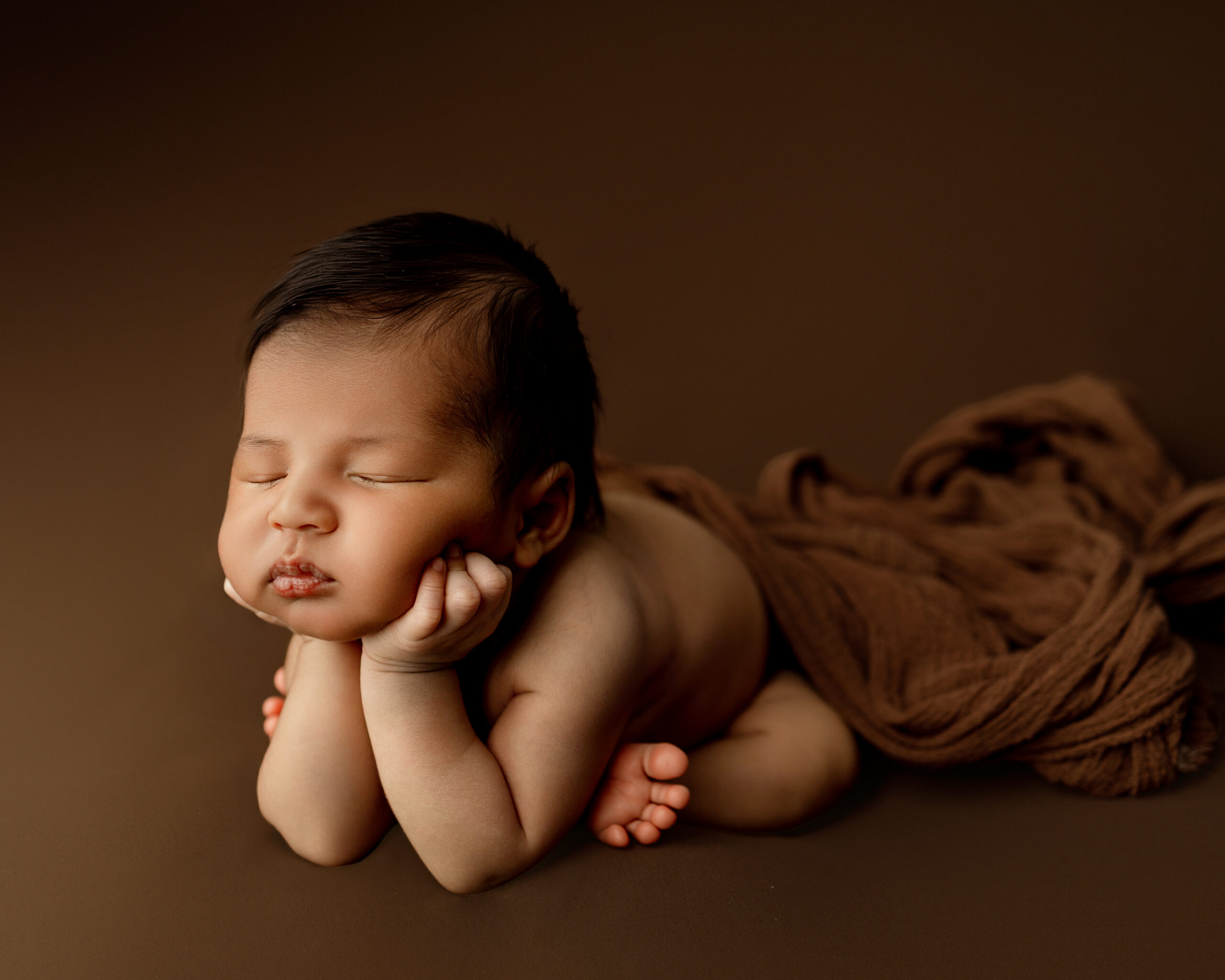 6 week old baby in newborn session in Yucaipa newborn studio, posed with chin resting on tiny hands on a brown backdrop.