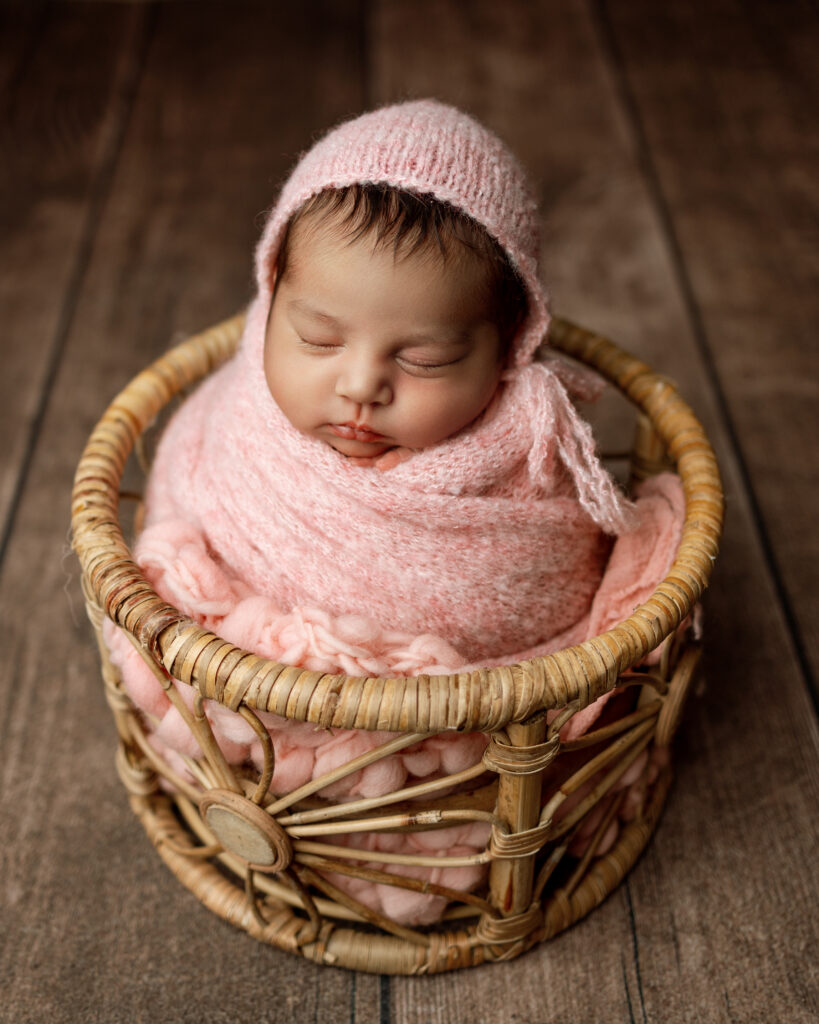 Sleeping baby girl wrapped in a soft pink knit bonnet and blanket in a wicker basket by an Inland Empire newborn photographer.