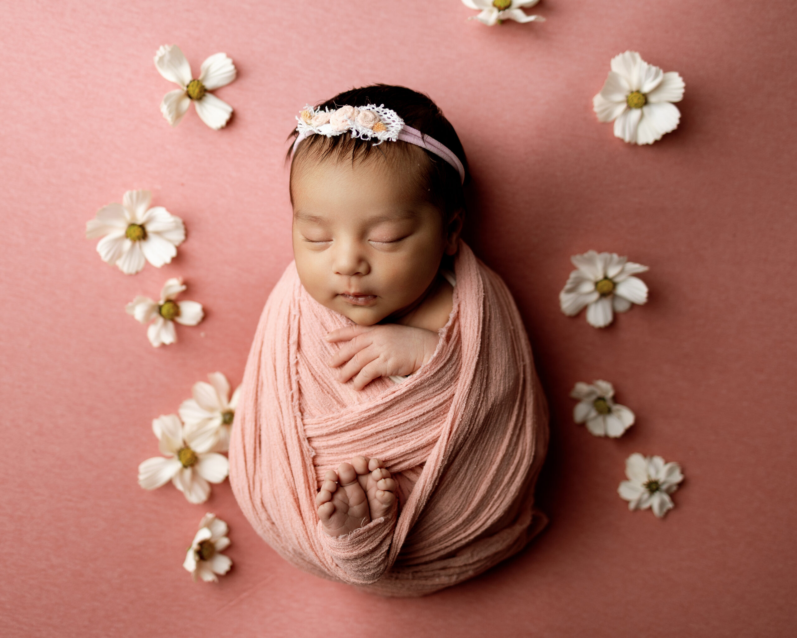 Sleeping newborn wrapped in a soft pink swaddle surrounded by white flowers, featured in a blog about newborn growth spurts.