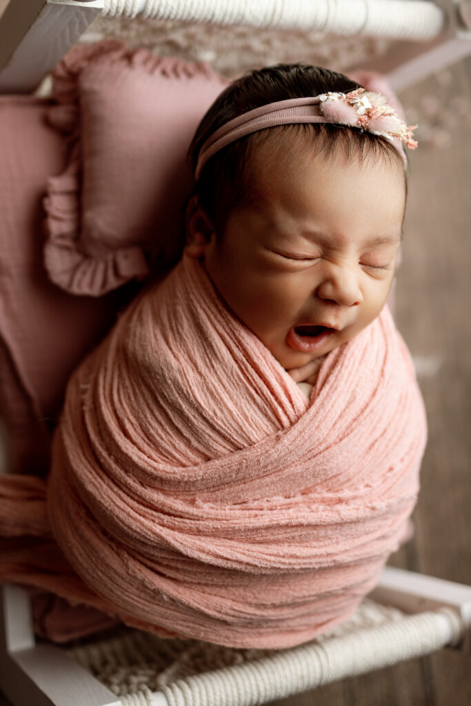 Yawning newborn baby in a blush swaddle resting on a miniature bed during a session with a newborn photographer in the Inland Empire.