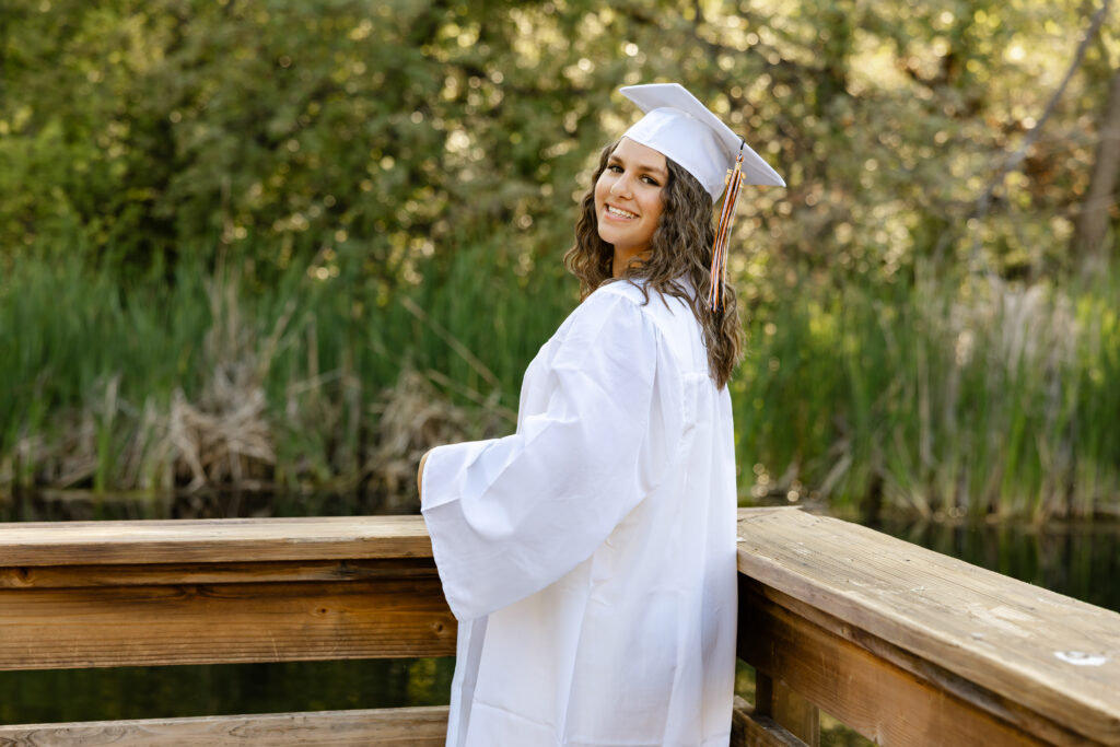 Yucaipa High School senior wearing a white cap and gown for graduation photos outdoors by a wooded pond.